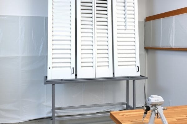 White plantation shutter panels drying on a rack in a workshop lined with plastic sheeting