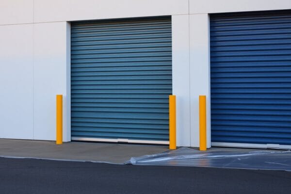Steel roll up doors on a storage building, one freshly painted deep blue, with yellow bollards