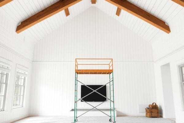 A vaulted white plank ceiling with stained wood beams and a rolling scaffold tower on a covered floor